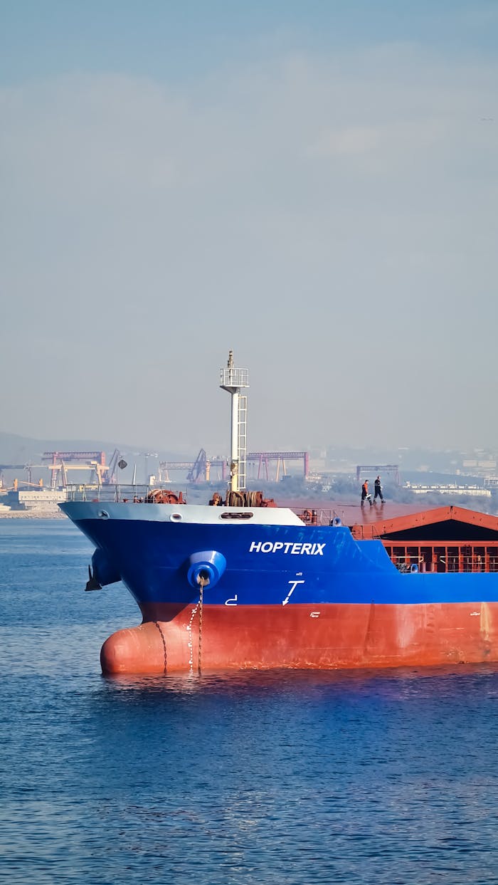 Close-up of a blue cargo ship on calm waters with an industrial port in the background.